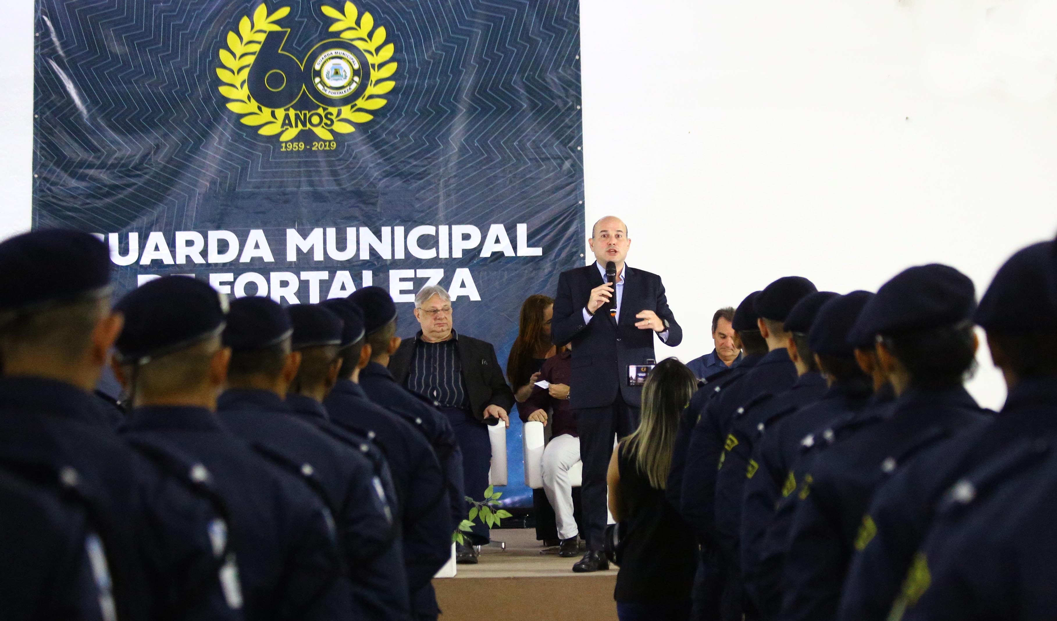 prefeito em palco falando ao microfone para guardas municipais em pé na frente